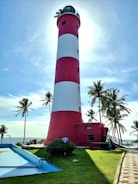 A tall, red and white striped lighthouse stands against a bright sky, surrounded by palm trees. The sun creates a halo effect around the top of the lighthouse, casting shadows on the green grassy area below. A small path leads towards the structure, and there are signs and a blue water feature nearby.