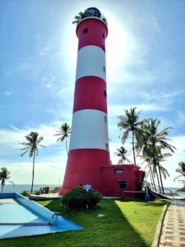 A tall, red and white striped lighthouse stands against a bright sky, surrounded by palm trees. The sun creates a halo effect around the top of the lighthouse, casting shadows on the green grassy area below. A small path leads towards the structure, and there are signs and a blue water feature nearby.