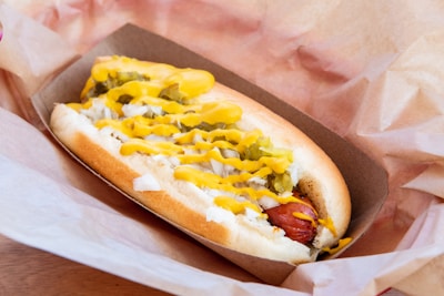 A hot dog nestled in a soft bun, topped with chopped onions, relish, and a generous drizzle of mustard. It is placed in a cardboard tray lined with crumpled paper.