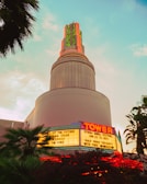 A vintage-style cinema building with neon signs featuring the word 'Tower'. The marquee lists several movie titles and events, illuminated against the backdrop of a pastel sky during twilight. The architecture is reminiscent of an Art Deco style, and palm trees surround the structure.