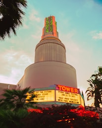 A vintage-style cinema building with neon signs featuring the word 'Tower'. The marquee lists several movie titles and events, illuminated against the backdrop of a pastel sky during twilight. The architecture is reminiscent of an Art Deco style, and palm trees surround the structure.