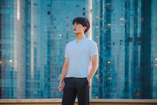 A person stands confidently in front of a backdrop of modern, glass skyscrapers. The individual is wearing a white shirt and dark pants, looking upwards with a contemplative expression.