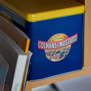 A vintage-style tin container with a blue body and a bright yellow lid. The container features a large label with the text 'Colman's Mustard' and an illustration that includes factories under a yellow sky. Several books are partially visible beside the tin, which appears to be situated on a wooden shelf.