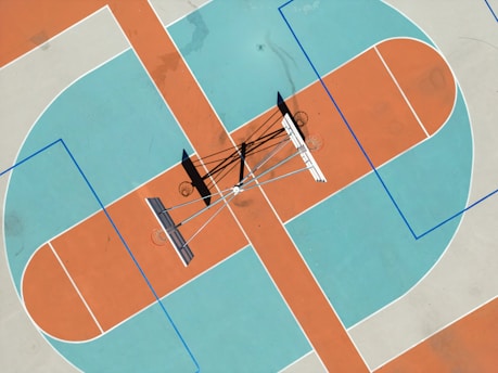 An aerial view of a basketball court with distinctive orange and blue sections. Multiple basketball hoops are positioned at different angles, casting shadows on the ground.