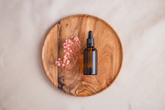 A brown glass dropper bottle is placed on a round wooden plate, accompanied by delicate pink dried flowers. The background is a neutral beige fabric.
