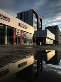 A modern urban setting with tall buildings reflecting on a wet surface after rain. The glass facade of the buildings catches the late afternoon sunlight, creating a striking contrast with the dark clouds above. The area appears quiet and empty, adding to the sleek, polished look of the architecture.