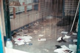 A cluttered and dusty room with scattered papers and debris on the floor. A shelf on the left contains folded textiles and a red container. The overall scene feels neglected with dirty windows and disorganized items.