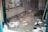 A cluttered and dusty room with scattered papers and debris on the floor. A shelf on the left contains folded textiles and a red container. The overall scene feels neglected with dirty windows and disorganized items.