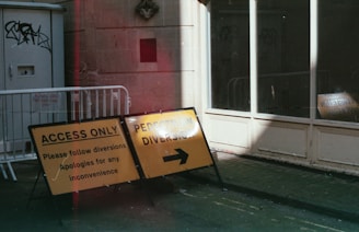 Yellow road signs with black text indicate 'Access Only' and 'Pedestrian Diversion' with arrows pointing to the right. Metal barricades are situated next to the signs, positioned on a street in front of a building with large windows and a door. Graffiti is visible on the door to the left. Partial sunlight illuminates parts of the scene, creating areas of shadow and light.