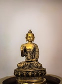 A golden statue of Buddha sitting cross-legged with an intricate design, holding one hand up in a gesture of teaching or reassurance. The sculpture features detailed engravings and rests on a decorated pedestal, with a simple neutral background that highlights its elegance.