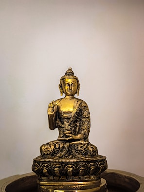 A golden statue of Buddha sitting cross-legged with an intricate design, holding one hand up in a gesture of teaching or reassurance. The sculpture features detailed engravings and rests on a decorated pedestal, with a simple neutral background that highlights its elegance.