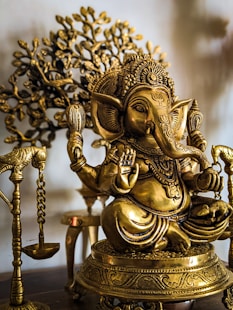 A detailed brass statue of the Hindu deity Ganesha, adorned with intricate carvings and ornaments. The statue is seated and surrounded by other brass elements, with a decorative tree in the background.