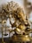 A detailed brass statue of the Hindu deity Ganesha, adorned with intricate carvings and ornaments. The statue is seated and surrounded by other brass elements, with a decorative tree in the background.