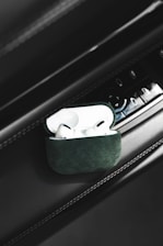 A pair of white wireless earbuds placed inside a green case are positioned on a car's interior panel. There are various buttons and controls visible on the panel, suggesting it is part of a car door or dashboard.
