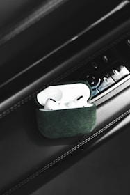 A pair of white wireless earbuds placed inside a green case are positioned on a car's interior panel. There are various buttons and controls visible on the panel, suggesting it is part of a car door or dashboard.