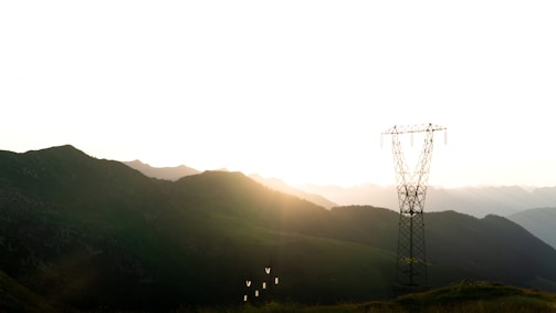 A transmission tower stands against the backdrop of a mountainous landscape at sunset. The sun is partially hidden behind the mountains, casting a warm glow over the scene. The sky is bright and clear, while the dark silhouette of the mountains adds contrast.