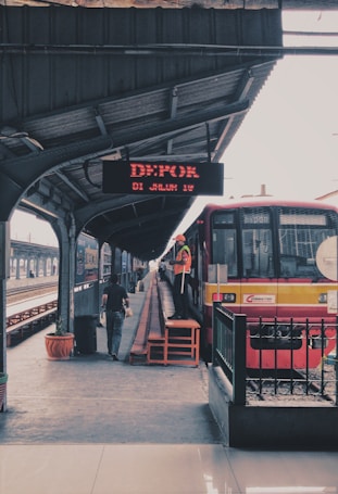 A train platform with a red and yellow train stopped at a station. A digital display board above indicates destination information. One person is walking alongside the train, and another person in a safety vest is standing on a small platform beside the train. The station has a metal roof and appears to be relatively empty with a planted pot under the roof structure.