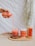 A hand pours a pink beverage into a clear glass filled with ice cubes. The scene includes a woven mat with two bottles, one labeled 'Sequoia' in orange and white, and an additional cup with the same label. The background features textured white fabric and hanging greenery.