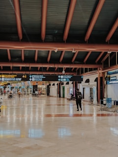 A spacious airport terminal with a polished floor and high ceiling supported by large beams. Overhead signs provide directions to various airport facilities such as departure gates and medical assistance. Few travelers are present, including one person walking with luggage.