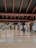 A spacious airport terminal with a polished floor and high ceiling supported by large beams. Overhead signs provide directions to various airport facilities such as departure gates and medical assistance. Few travelers are present, including one person walking with luggage.