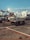A tarmac scene at an airport with service vehicles parked. The foreground features a luggage cart with equipment on it, while the background shows several airplanes parked at the terminal. The lighting creates a calm, early evening ambiance with a clear sky.