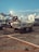 A tarmac scene at an airport with service vehicles parked. The foreground features a luggage cart with equipment on it, while the background shows several airplanes parked at the terminal. The lighting creates a calm, early evening ambiance with a clear sky.