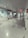 A clean, empty hallway with glossy tiled flooring and white walls. Overhead, there is a ceiling with recessed lighting, and on the right side, a series of doors and signs are visible. There is a television mounted on the wall and some signage indicating directions or information.