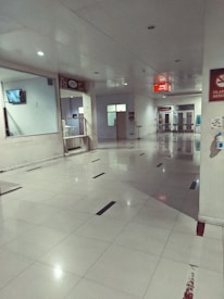 A clean, empty hallway with glossy tiled flooring and white walls. Overhead, there is a ceiling with recessed lighting, and on the right side, a series of doors and signs are visible. There is a television mounted on the wall and some signage indicating directions or information.