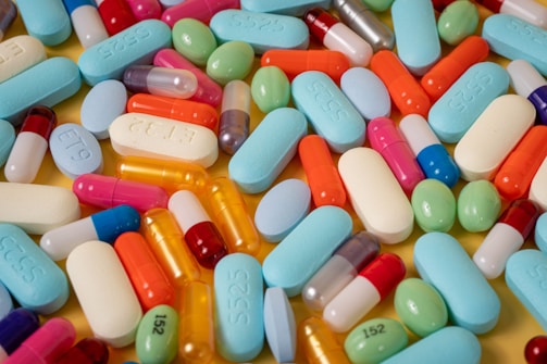A variety of colorful pills and capsules are scattered across a surface. The colors range from blue, green, orange, to pink, and there are different shapes including oval, round, and cylindrical capsules.