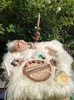 A spiritual altar is set up outdoors on a fluffy white surface with various crystals, a wooden board spelled 'ENERGIES' with wooden letters, tarot or oracle cards, a candle, and a bowl of pink Himalayan salt. Green foliage surrounds the setup, creating a serene and natural backdrop.