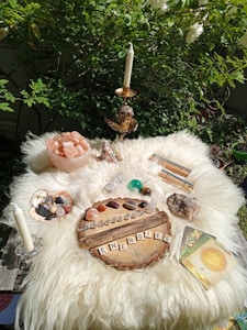 A spiritual altar is set up outdoors on a fluffy white surface with various crystals, a wooden board spelled 'ENERGIES' with wooden letters, tarot or oracle cards, a candle, and a bowl of pink Himalayan salt. Green foliage surrounds the setup, creating a serene and natural backdrop.