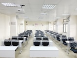 A classroom with rows of black chairs and white desks facing the front. The room is brightly lit with overhead lights and large windows on the right side. A projector is mounted on the ceiling, and there are decorations on the walls. The arrangement of furniture suggests a formal educational setting.
