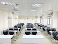 A classroom with rows of black chairs and white desks facing the front. The room is brightly lit with overhead lights and large windows on the right side. A projector is mounted on the ceiling, and there are decorations on the walls. The arrangement of furniture suggests a formal educational setting.