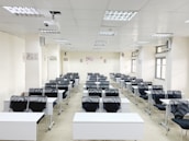 A classroom with rows of black chairs and white desks facing the front. The room is brightly lit with overhead lights and large windows on the right side. A projector is mounted on the ceiling, and there are decorations on the walls. The arrangement of furniture suggests a formal educational setting.