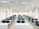 A classroom with rows of black chairs and white desks facing the front. The room is brightly lit with overhead lights and large windows on the right side. A projector is mounted on the ceiling, and there are decorations on the walls. The arrangement of furniture suggests a formal educational setting.