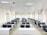 A classroom with rows of black chairs and white desks facing the front. The room is brightly lit with overhead lights and large windows on the right side. A projector is mounted on the ceiling, and there are decorations on the walls. The arrangement of furniture suggests a formal educational setting.