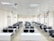 A classroom with rows of black chairs and white desks facing the front. The room is brightly lit with overhead lights and large windows on the right side. A projector is mounted on the ceiling, and there are decorations on the walls. The arrangement of furniture suggests a formal educational setting.