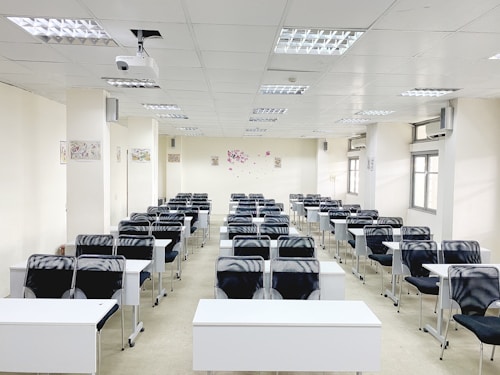 A classroom with rows of black chairs and white desks facing the front. The room is brightly lit with overhead lights and large windows on the right side. A projector is mounted on the ceiling, and there are decorations on the walls. The arrangement of furniture suggests a formal educational setting.