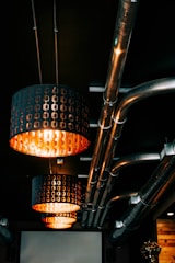Overhead lighting fixtures with an industrial design hang from the ceiling, casting a warm orange glow. The ceiling features several exposed metal pipes, giving the space a modern yet utilitarian look. The lighting fixtures have a perforated pattern allowing the light to shine through, enhancing the ambiance.