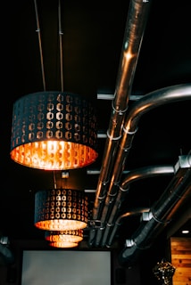 Overhead lighting fixtures with an industrial design hang from the ceiling, casting a warm orange glow. The ceiling features several exposed metal pipes, giving the space a modern yet utilitarian look. The lighting fixtures have a perforated pattern allowing the light to shine through, enhancing the ambiance.