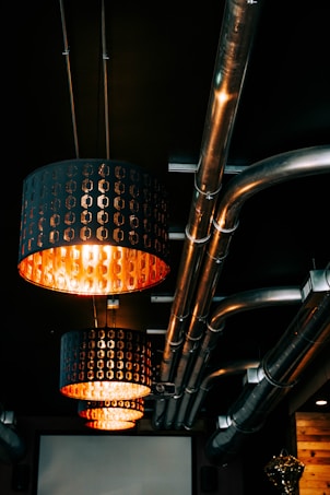 Overhead lighting fixtures with an industrial design hang from the ceiling, casting a warm orange glow. The ceiling features several exposed metal pipes, giving the space a modern yet utilitarian look. The lighting fixtures have a perforated pattern allowing the light to shine through, enhancing the ambiance.