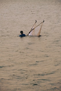 A person standing waist-deep in water holds a fishing net that is supported by two poles. The surface of the water is calm with gentle ripples, and the lighting suggests it may be early morning or late afternoon.