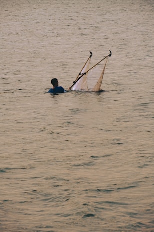 A person standing waist-deep in water holds a fishing net that is supported by two poles. The surface of the water is calm with gentle ripples, and the lighting suggests it may be early morning or late afternoon.