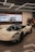A sleek, white high-performance sports car is showcased in a modern showroom. The vehicle has aerodynamic curves, tinted windows, and large black wheels. Above the car, there are suspended geometric light fixtures. In the background, a large screen displays images and text, likely detailing automotive history or specifications.