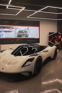 A sleek, white high-performance sports car is showcased in a modern showroom. The vehicle has aerodynamic curves, tinted windows, and large black wheels. Above the car, there are suspended geometric light fixtures. In the background, a large screen displays images and text, likely detailing automotive history or specifications.