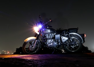 A motorcycle is parked on a rough terrain during nighttime. The vehicle is illuminated by a bright headlight, while the background fades into darkness with a distant cityscape visible. The motorcycle appears to be a Royal Enfield, showcasing its classic design.