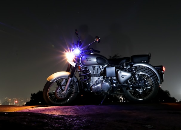 A motorcycle is parked on a rough terrain during nighttime. The vehicle is illuminated by a bright headlight, while the background fades into darkness with a distant cityscape visible. The motorcycle appears to be a Royal Enfield, showcasing its classic design.