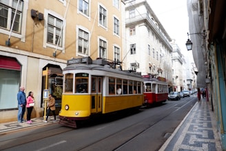 A traditional yellow tram labeled 'Prazeres 28E' travels down a narrow street lined with historic buildings. A red tram follows closely behind. Several pedestrians stand on the sidewalk, some waiting at a tram stop post. The street is flanked by ornate architecture typical of European urban landscapes.