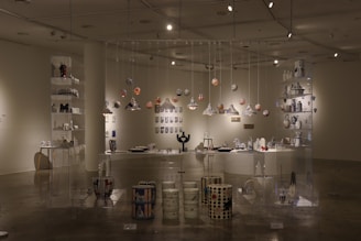 An art gallery space with a variety of ceramic and pottery pieces displayed on shelves and hanging from the ceiling. The room features well-lit, glass shelving units containing colorful and intricately designed vases, bowls, and decorative items. The atmosphere is enhanced by strategically placed spotlights, creating shadows and highlights that accentuate the art. The overall arrangement suggests a thoughtful and aesthetically pleasing display meant to showcase the creativity and craftsmanship of the pieces.
