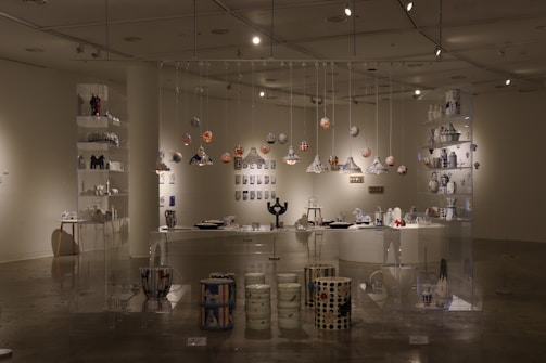 An art gallery space with a variety of ceramic and pottery pieces displayed on shelves and hanging from the ceiling. The room features well-lit, glass shelving units containing colorful and intricately designed vases, bowls, and decorative items. The atmosphere is enhanced by strategically placed spotlights, creating shadows and highlights that accentuate the art. The overall arrangement suggests a thoughtful and aesthetically pleasing display meant to showcase the creativity and craftsmanship of the pieces.